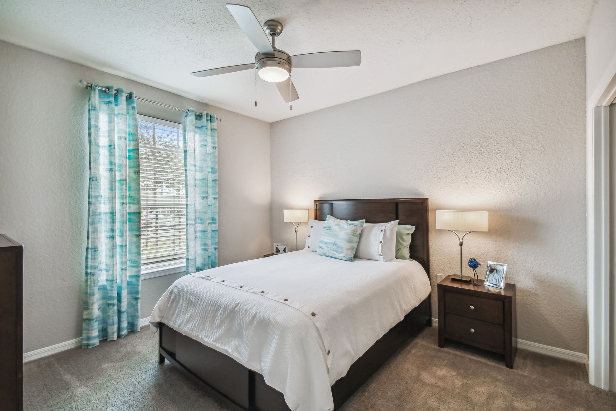 3 Bedroom Apartments in Brandon, FL - 211 Grand Isle - a Bedroom with Plush Carpeting, a Bed with White Bedding and a Brown Wood Bedframe, Two Bedside Tables with Drawers and Lamps, a Large Wood Dresser, a Ceiling Fan with Lighting, and a Large Window with White Blinds and Light Blue Pattern Curtains