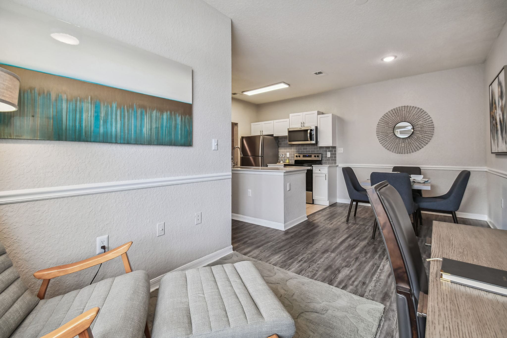 **Alt Text Description:** View of a contemporary open-concept living space showcasing a reading nook, kitchen, and dining area. In the foreground, a mid-century modern lounge chair with light gray upholstery and wooden arms is paired with a matching ottoman. To the right, part of a wooden desk and a dark leather chair are visible, suggesting a compact workspace. A wide piece of abstract wall art in shades of teal, brown, and white adds a pop of color. The kitchen features white cabinetry, stainless steel appliances, gray subway tile backsplash, and a quartz countertop island with a built-in sink. Beyond the kitchen is a dining area with a square table, dark upholstered chairs, a decorative wire mirror, and abstract artwork on the wall. The space is unified by wood-style flooring and soft, neutral wall tones, creating a cozy, modern atmosphere. Modern open-concept interior featuring a small reading nook, kitchen, and dining area. In the foreground, there is a mid-century style lounge chair with a matching ottoman upholstered in light gray fabric. A piece of abstract wall art in teal and bronze tones hangs above the chair. To the right, part of a wooden desk with a notebook and a dark leather chair is visible. The space opens into a kitchen with white cabinetry, stainless steel appliances, gray subway tile backsplash, and a peninsula with an overhang for seating. Adjacent to the kitchen is a contemporary dining area with a square table, set for four, surrounded by upholstered dark blue chairs. A modern wire-framed mirror and abstract artwork add decorative flair to the dining wall. The flooring alternates between wood-style laminate and tile, and soft, neutral wall tones unify the space.