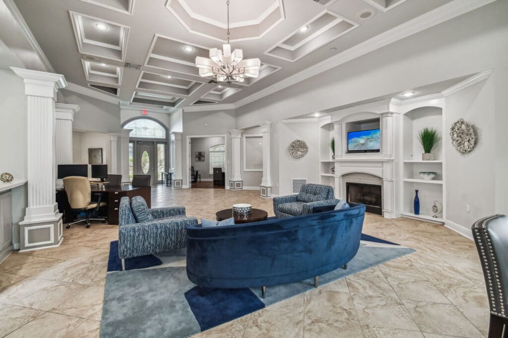 **Alt Text Description:** Elegant clubhouse lounge featuring a grand coffered ceiling with recessed lighting and a modern chandelier. The space includes a stylish seating area with a curved navy velvet sofa, two patterned armchairs, and a round wooden coffee table placed on a geometric area rug. A built-in white entertainment center with display shelves and a mounted flat-screen TV frames a central fireplace. Decorative accents include vases, plants, and sculptural wall pieces. To the left, a work or business center area is furnished with desks and office chairs, while the arched double front doors and additional rooms are visible in the background. The floor is finished with beige marble-look tile, and the overall design blends sophistication with comfort.