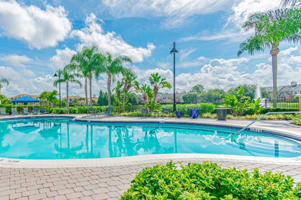This image showcases a **beautiful resort-style swimming pool** set within a tropical residential community. The clear blue pool water glistens under a bright sky dotted with fluffy white clouds. Surrounding the pool are **lush tropical plants** and **palm trees**, enhancing the serene, vacation-like atmosphere. The pool area is bordered by a **paved pool deck** with plenty of **lounge chairs** and **umbrella-covered seating areas** for relaxation. Decorative **blue planters** add a touch of color to the landscaping, and a **fountain in a nearby lake** creates a tranquil focal point in the background. Residential buildings with light-colored facades are visible beyond the pool area, suggesting this is part of a well-maintained apartment or condo community. Overall, the setting conveys a peaceful, upscale ambiance ideal for leisurely afternoons in the sun. Would you like a caption or promotional text for this image?