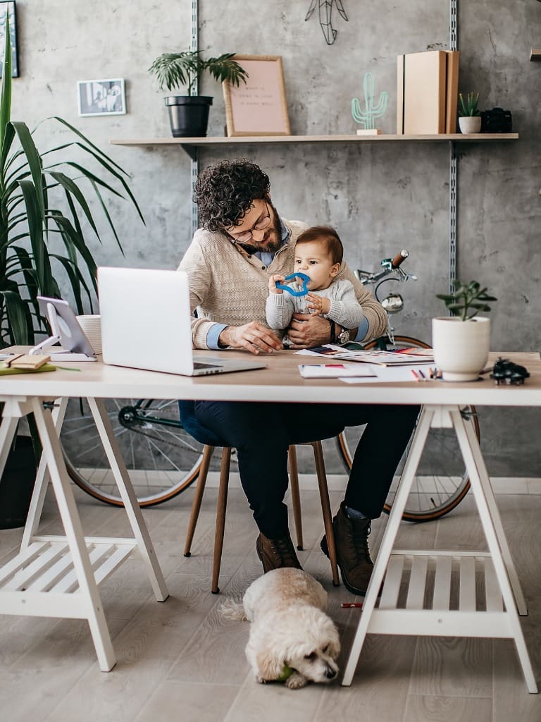 **Alt Text Description:** A father is working from home in a stylish modern office with concrete walls and minimalistic decor. He is seated at a wooden desk with a white trestle base, holding his baby on his lap while working on a laptop. The baby is playing with a blue toy, and both are dressed in cozy, neutral-toned sweaters. A small white dog lies under the desk, resting on the wooden floor. The room features green plants, a bicycle in the background, and wall-mounted shelves with books, a cactus lamp, and decorative items. The atmosphere is warm, productive, and family-oriented.