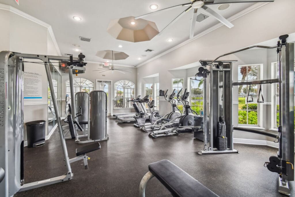 **Alt Text Description:** Modern fitness center filled with natural light from large, arched and rectangular windows that offer views of lush greenery outside. The gym is equipped with a variety of workout machines including ellipticals, treadmills, recumbent bikes, and multi-use strength training equipment. The space features high ceilings with stylish geometric light fixtures and oversized ceiling fans for ventilation. The interior is bright and clean, with neutral walls, black rubber flooring, and mirrored walls that enhance the sense of space.
